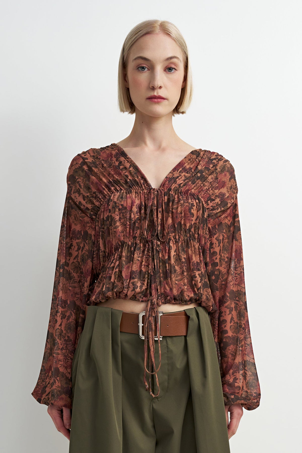 A model with a short blonde bob is wearing a long-sleeved, smocked, and tie-front blouse in a rust-brown and black abstract pattern, paired with olive green trousers and a brown belt, against a plain light gray background.