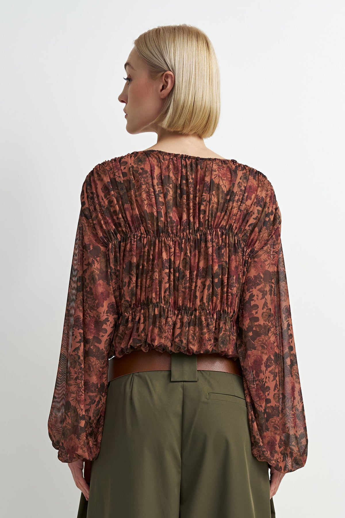 A woman shown from the back wearing a long-sleeve, gathered blouse with an abstract floral pattern in rust red, brown, and black, paired with olive green trousers against a light gray studio background.
