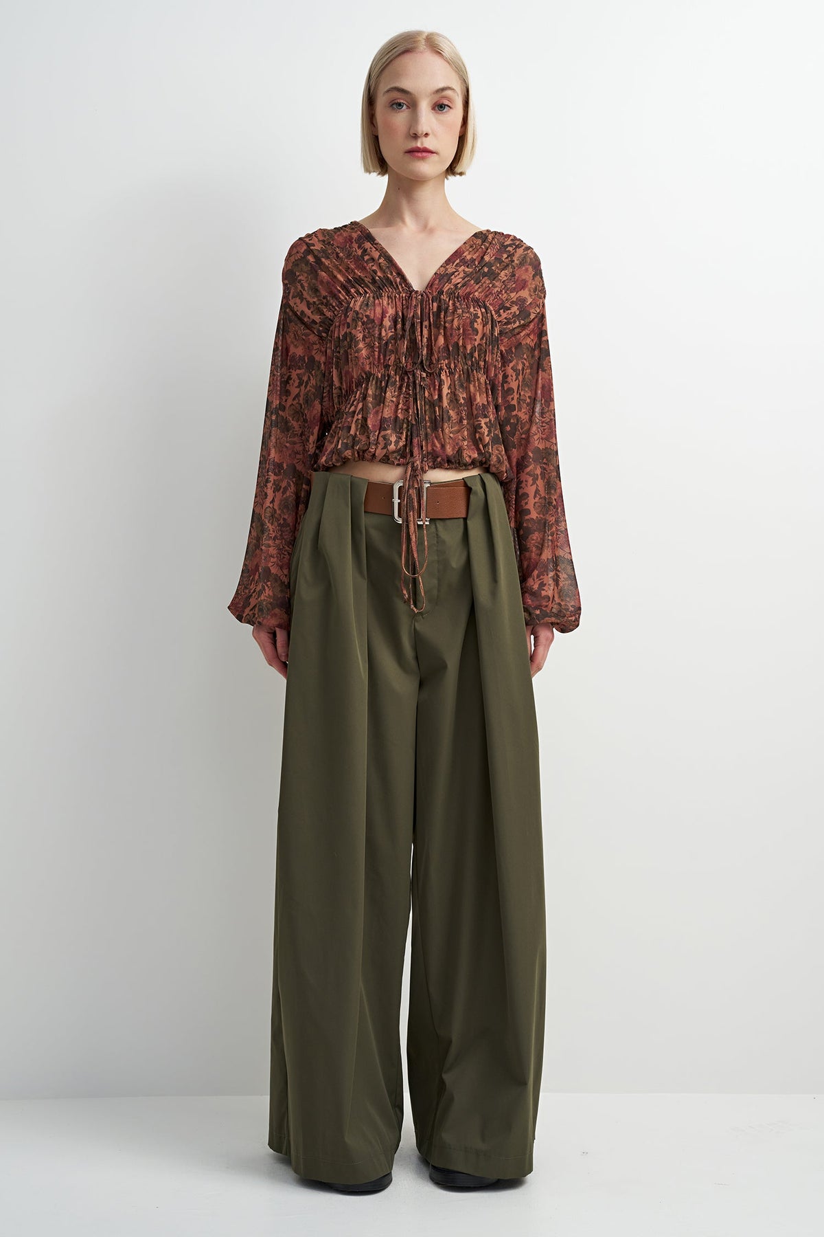 A model stands against a plain light gray studio background wearing a long-sleeve, gathered V-neck blouse with an abstract rust and brown pattern, paired with wide-leg olive green trousers and a brown belt.