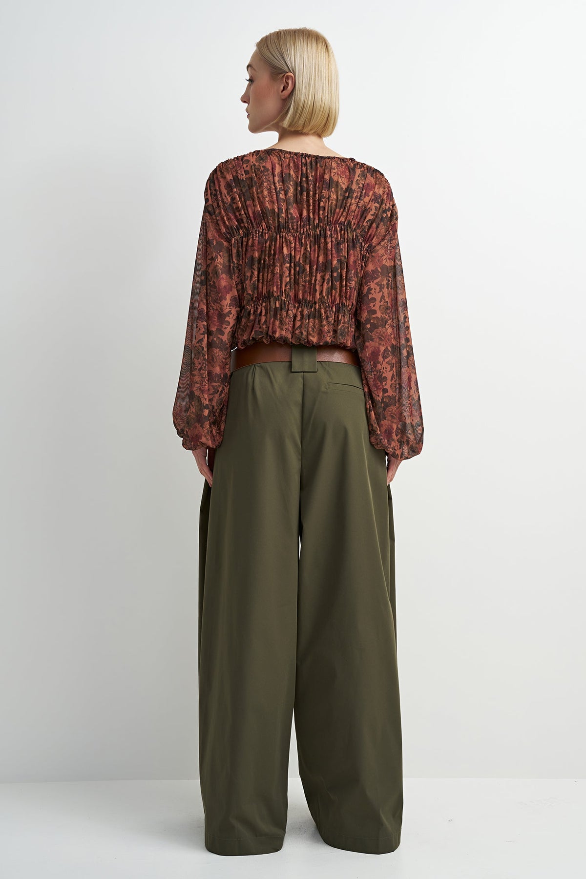 A model shown from the back wearing a long-sleeve, gathered, cropped blouse with a rust-red and black floral pattern, paired with olive green wide-leg trousers, against a plain light gray background.