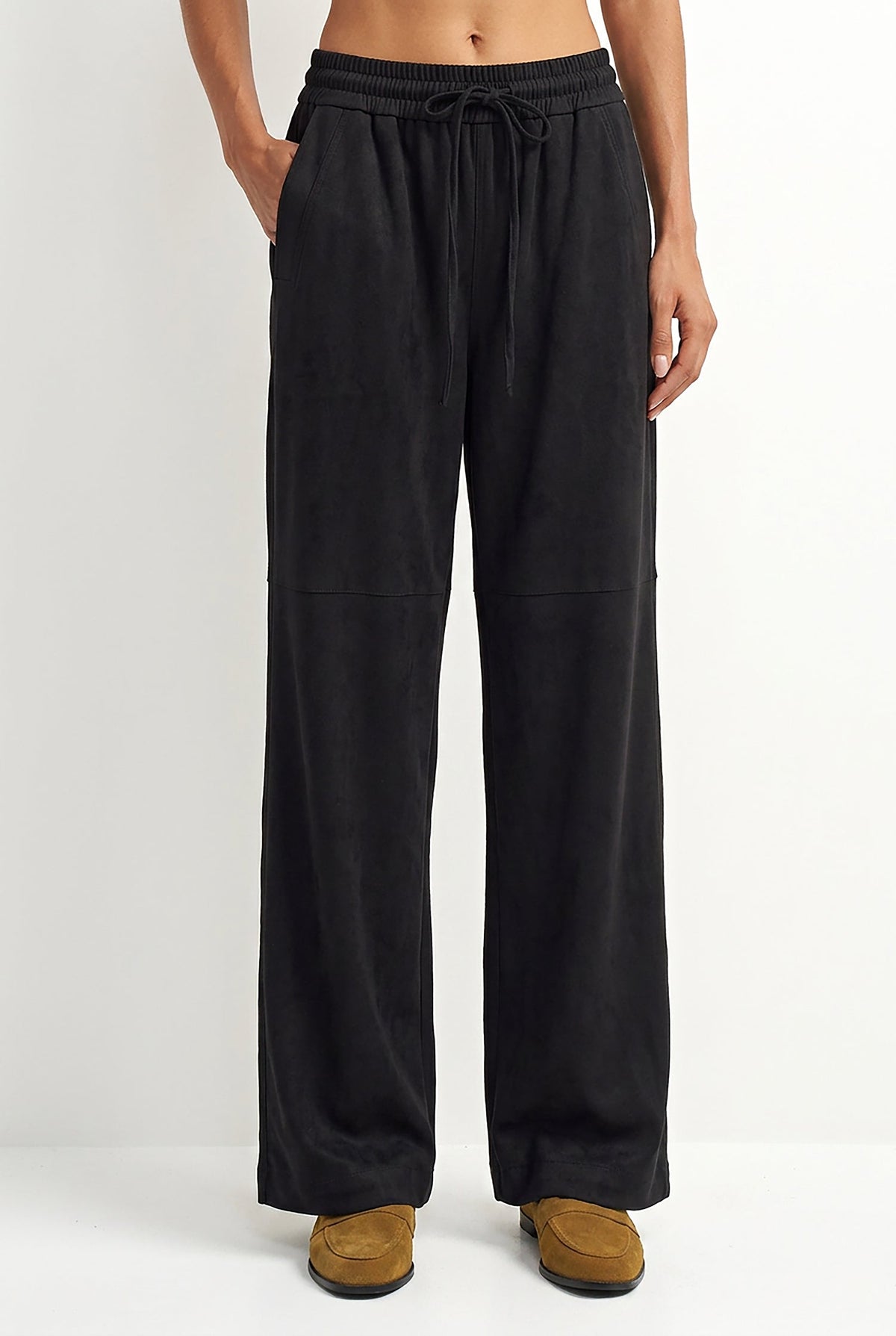 A person wearing black wide-leg drawstring pants with side pockets, paired with tan slip-on shoes, shown against a plain light gray studio background.