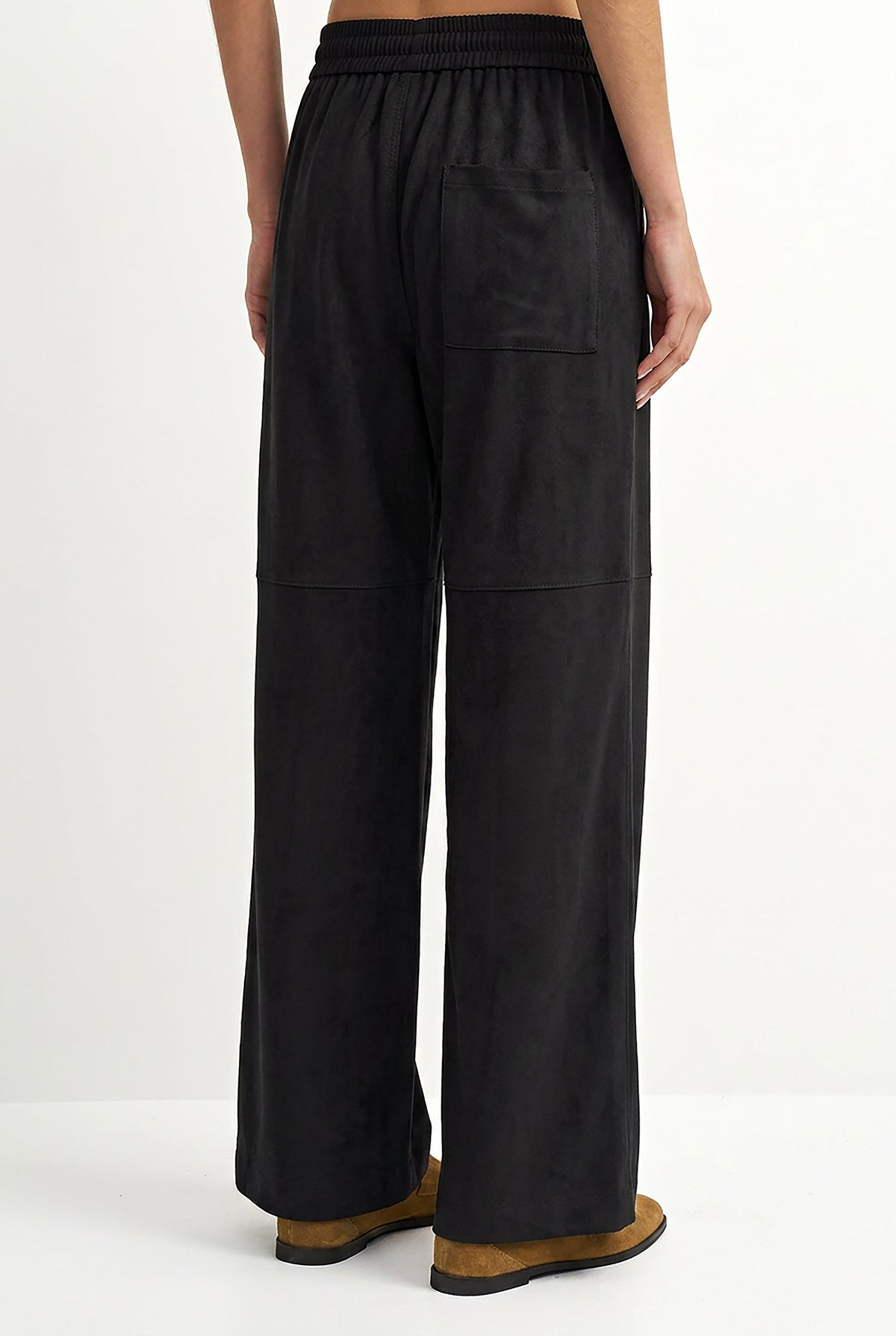 A person shown from the back wearing loose black wide-leg pants with an elastic waistband and brown shoes, photographed against a clean white studio background.
