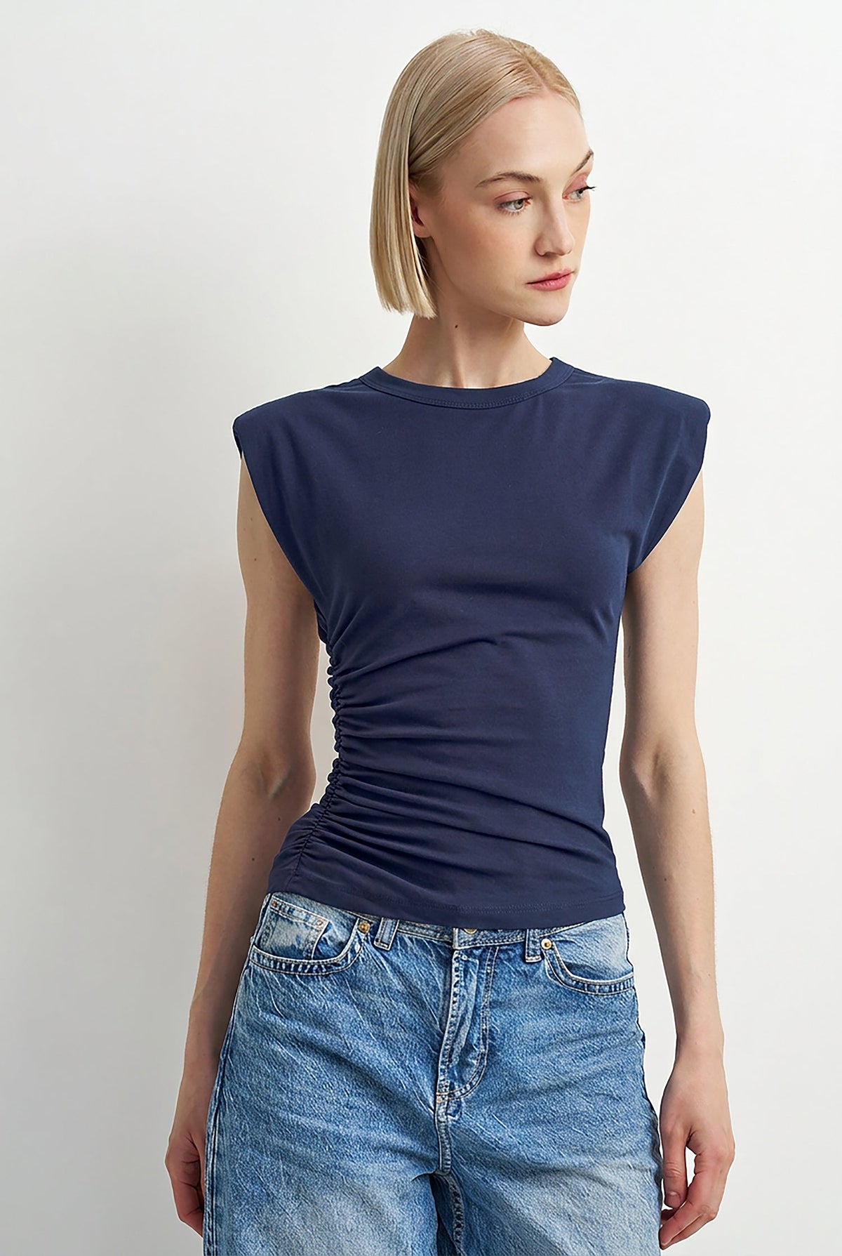 A woman modeling a navy blue sleeveless top with a ruched side detail, paired with light-wash blue jeans, against a plain light gray background.