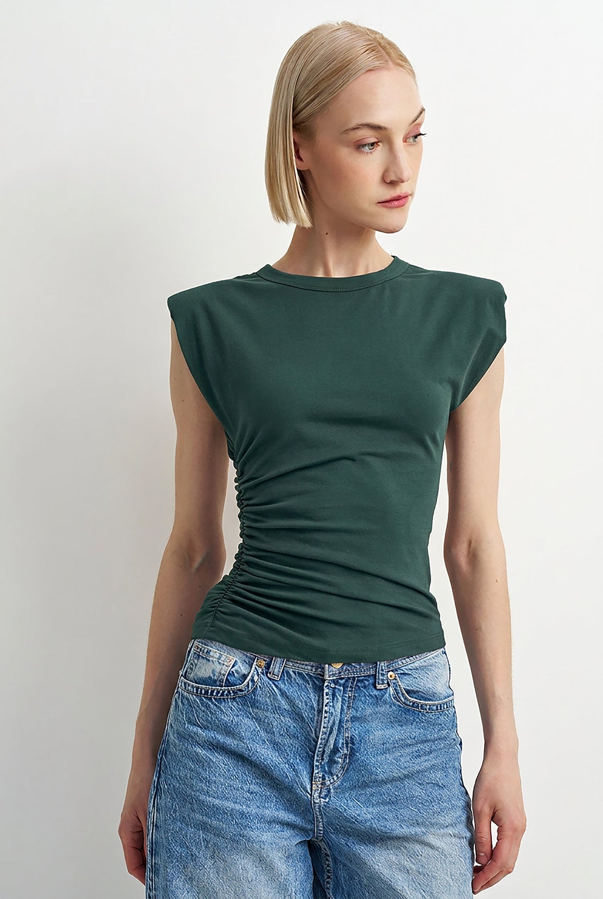 A woman modeling a dark green sleeveless top with ruching at the sides and blue jeans, photographed against a plain light gray studio background.