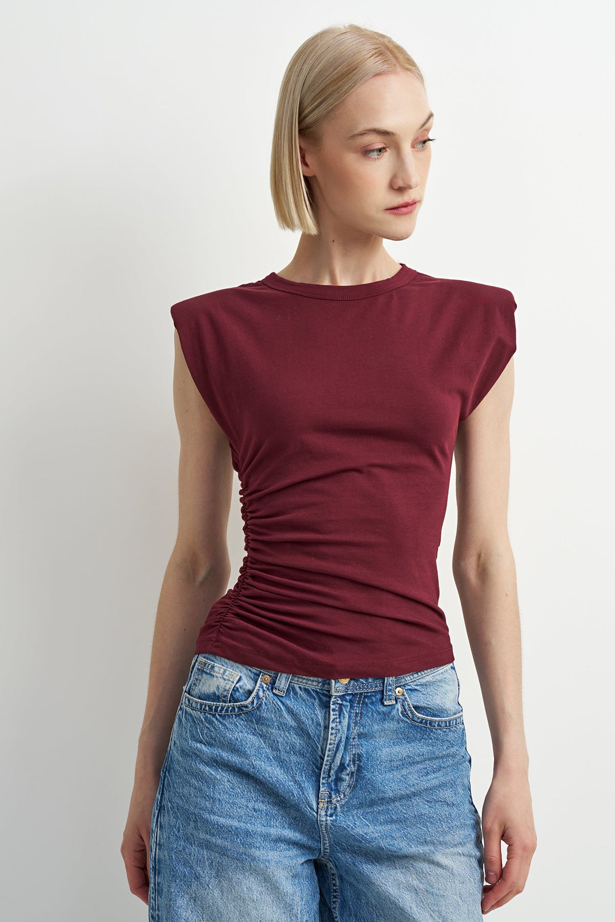 A woman wearing a sleeveless, ruched crewneck t-shirt in deep burgundy, paired with light wash blue jeans, photographed against a neutral light gray background.