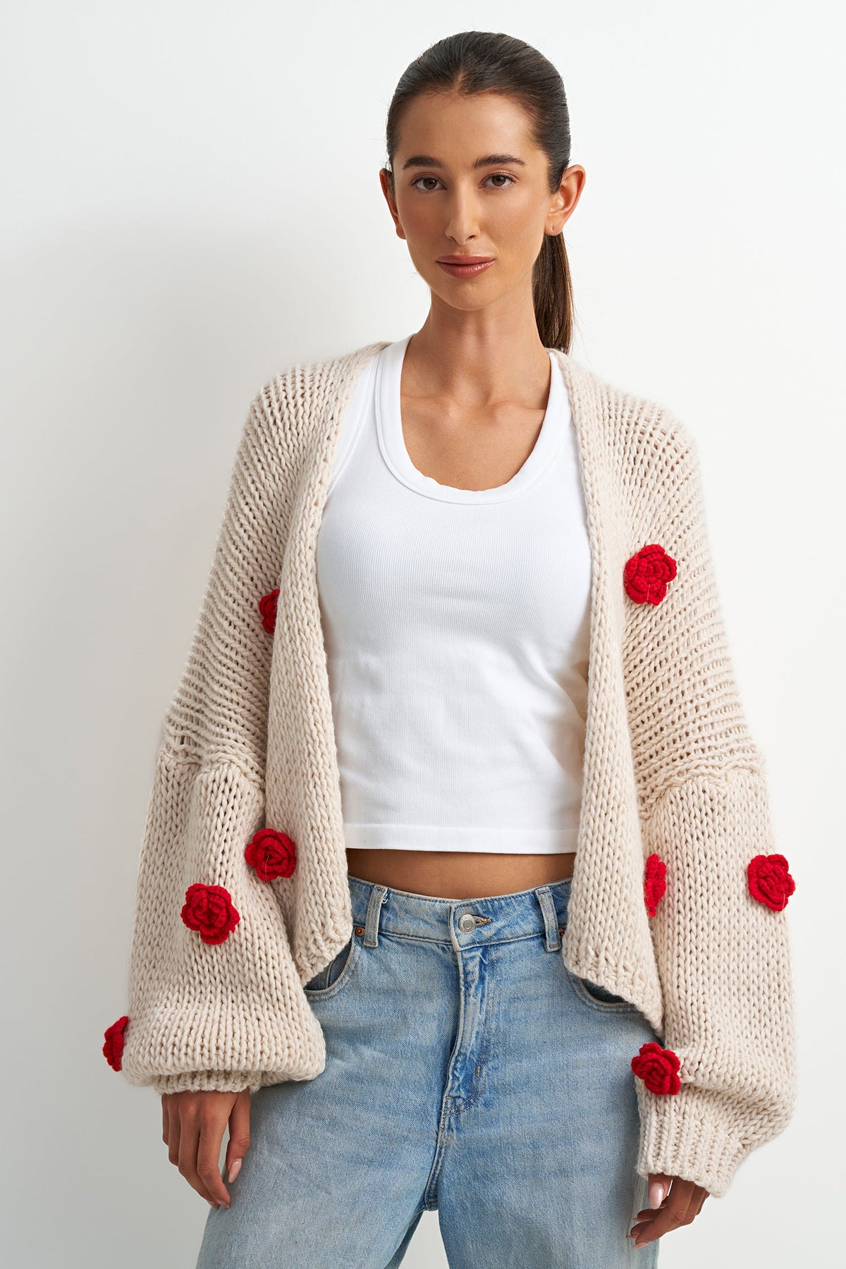 A woman modeling a cream-colored chunky knit cardigan with red pom-pom accents, worn over a white crop top and light blue jeans against a plain white background.