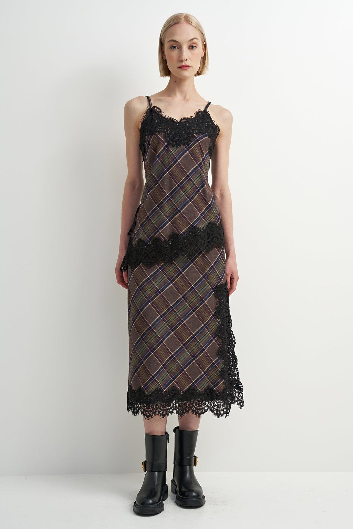A model wearing a sleeveless midi dress with a brown and black plaid pattern, black lace trim, and black ankle boots, standing against a plain white studio background.