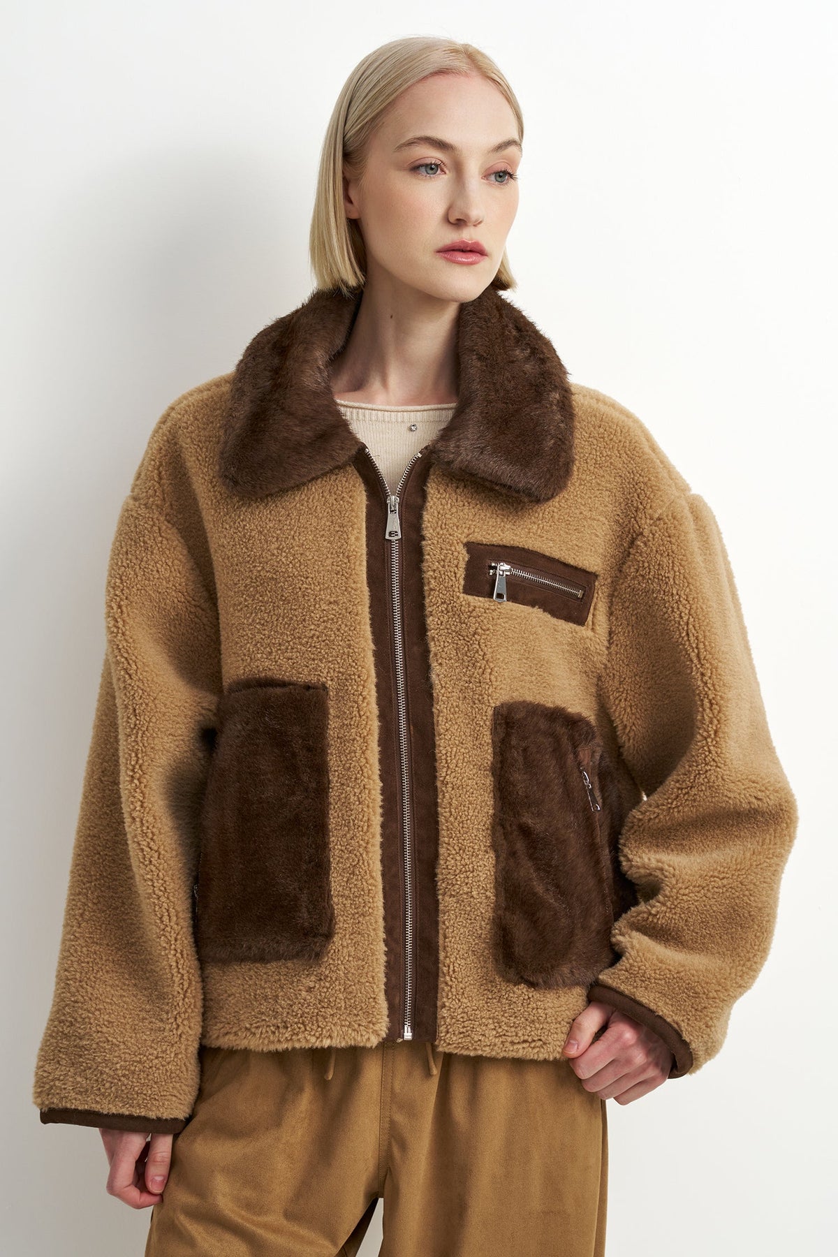 A model wearing a tan sherpa fleece jacket with dark brown faux-fur trim, a dark brown zipper, and two large dark brown patch pockets, paired with brown pants against a plain white studio background.