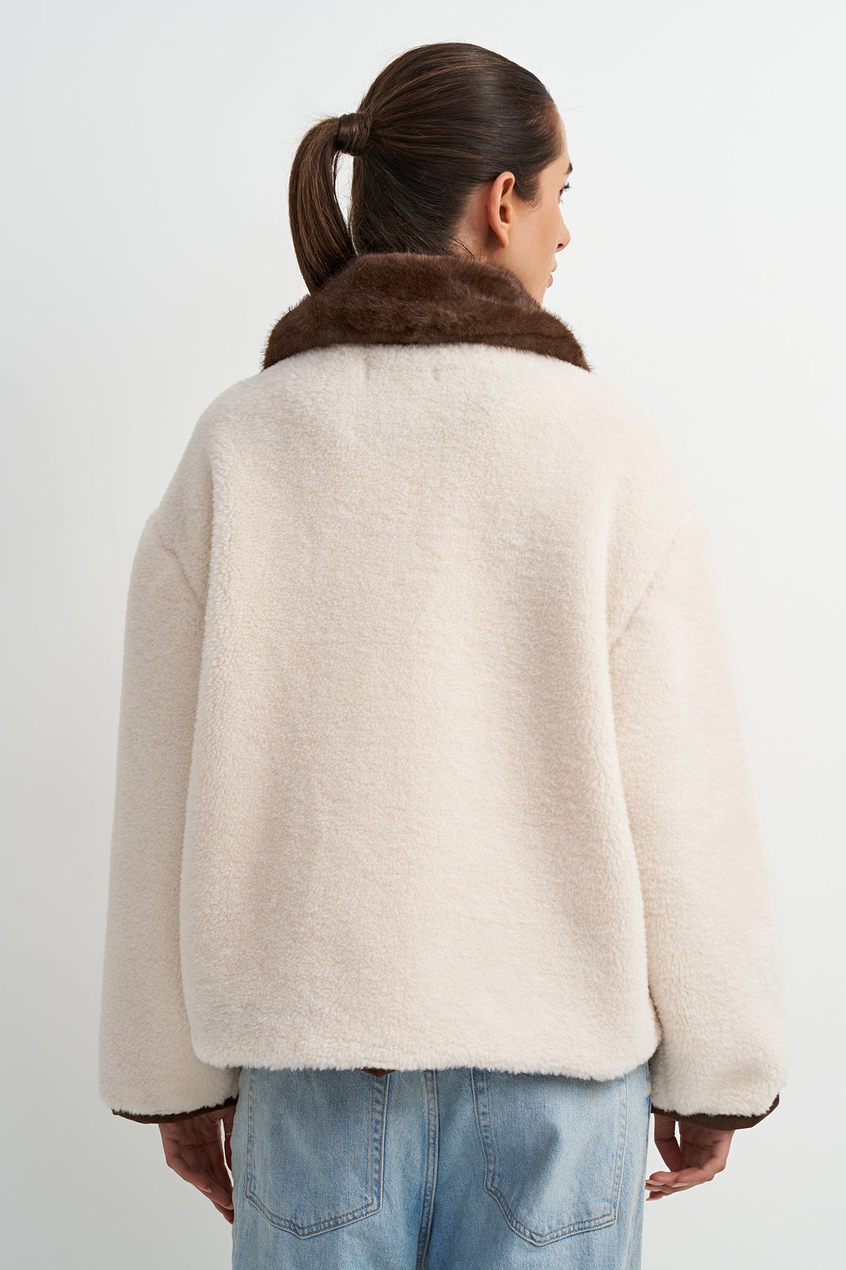 A woman shown from the back wearing a cream-colored fuzzy teddy jacket with a brown faux-fur collar, paired with light blue jeans, against a plain white studio background.