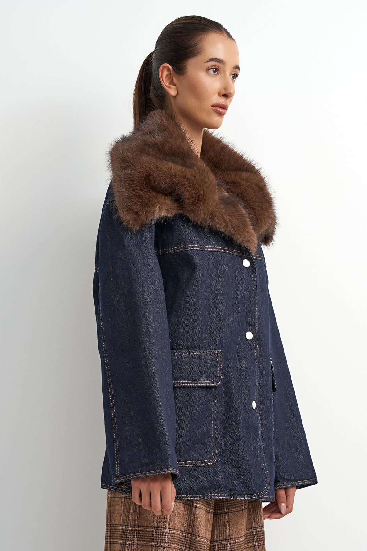 A woman wearing a dark blue denim jacket with a large brown fur collar, shown in profile against a plain light gray background.