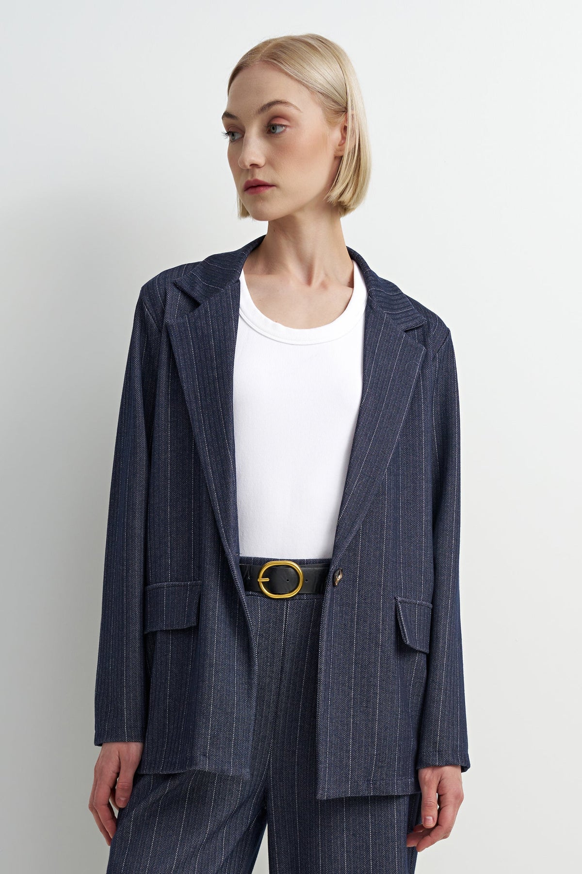 A woman modeling a navy blue pinstripe blazer with matching trousers, styled with a white crewneck top and a gold-buckled belt, against a plain light gray background.