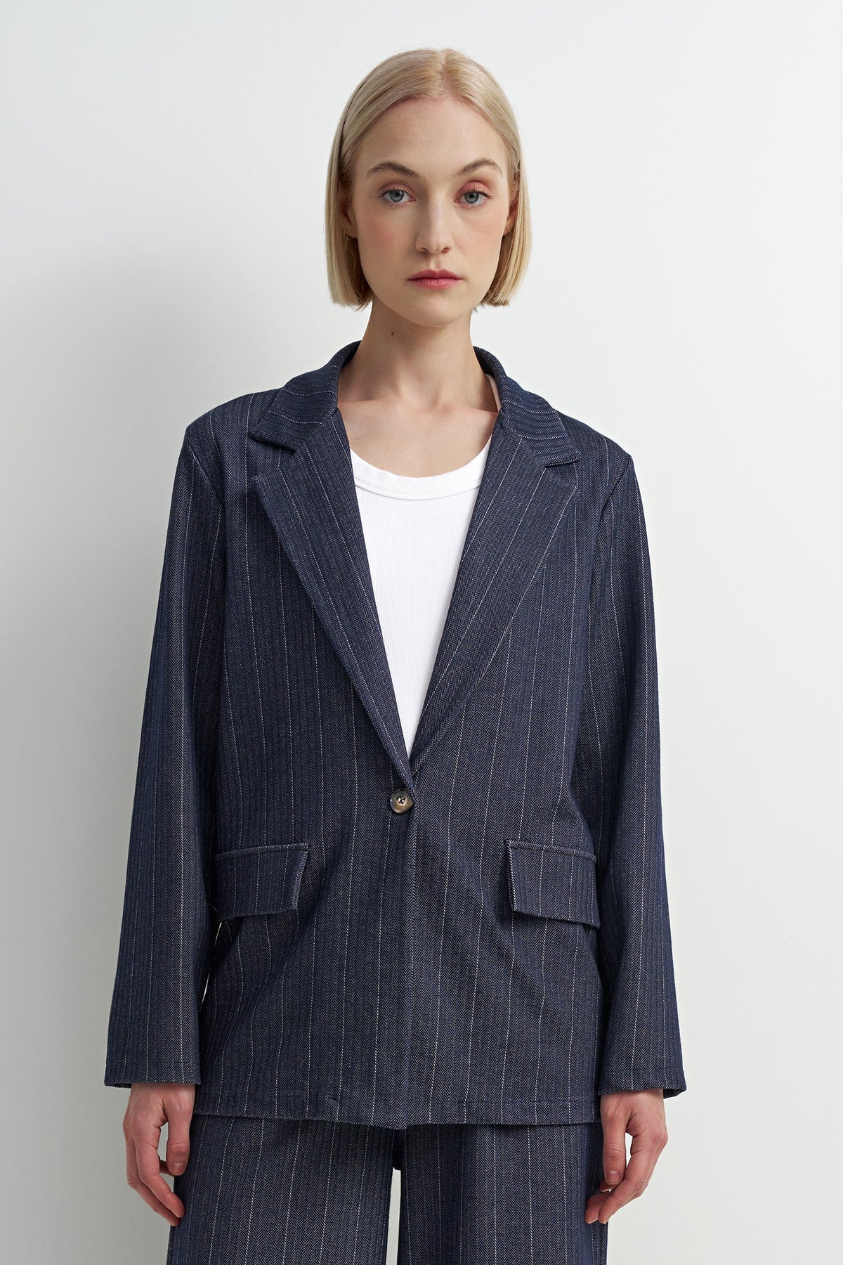 A woman modeling a navy blue pinstripe blazer with a single button closure, paired with a white crewneck t-shirt and matching navy pinstripe trousers, against a plain light gray background.