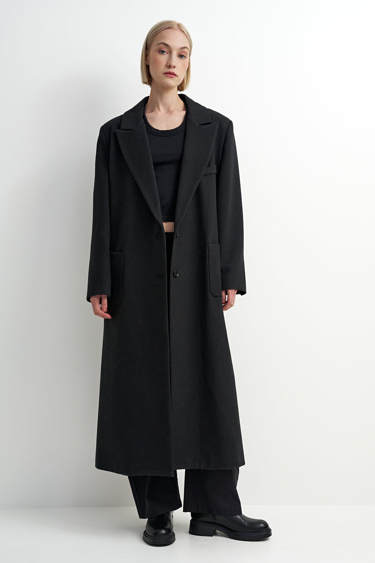 A model wearing a long black wool-blend coat over a black outfit, standing against a plain white studio background.