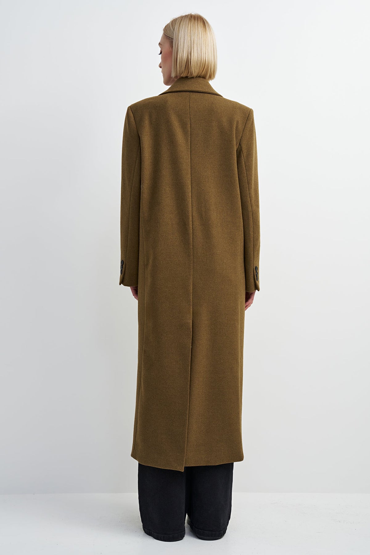 A model viewed from the back wearing a long, straight-line brown coat over black trousers, set against a plain light gray studio background.