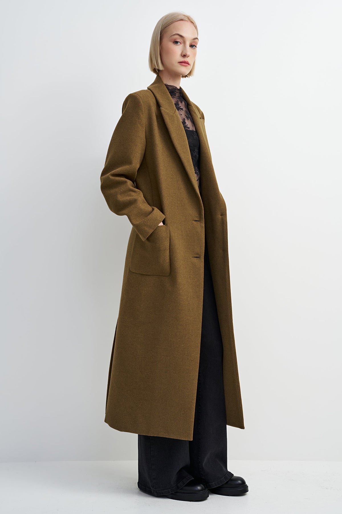 A woman modeling a long, mid-length olive-brown wool coat over a dark patterned top and black wide-leg trousers, standing against a plain light gray studio background.