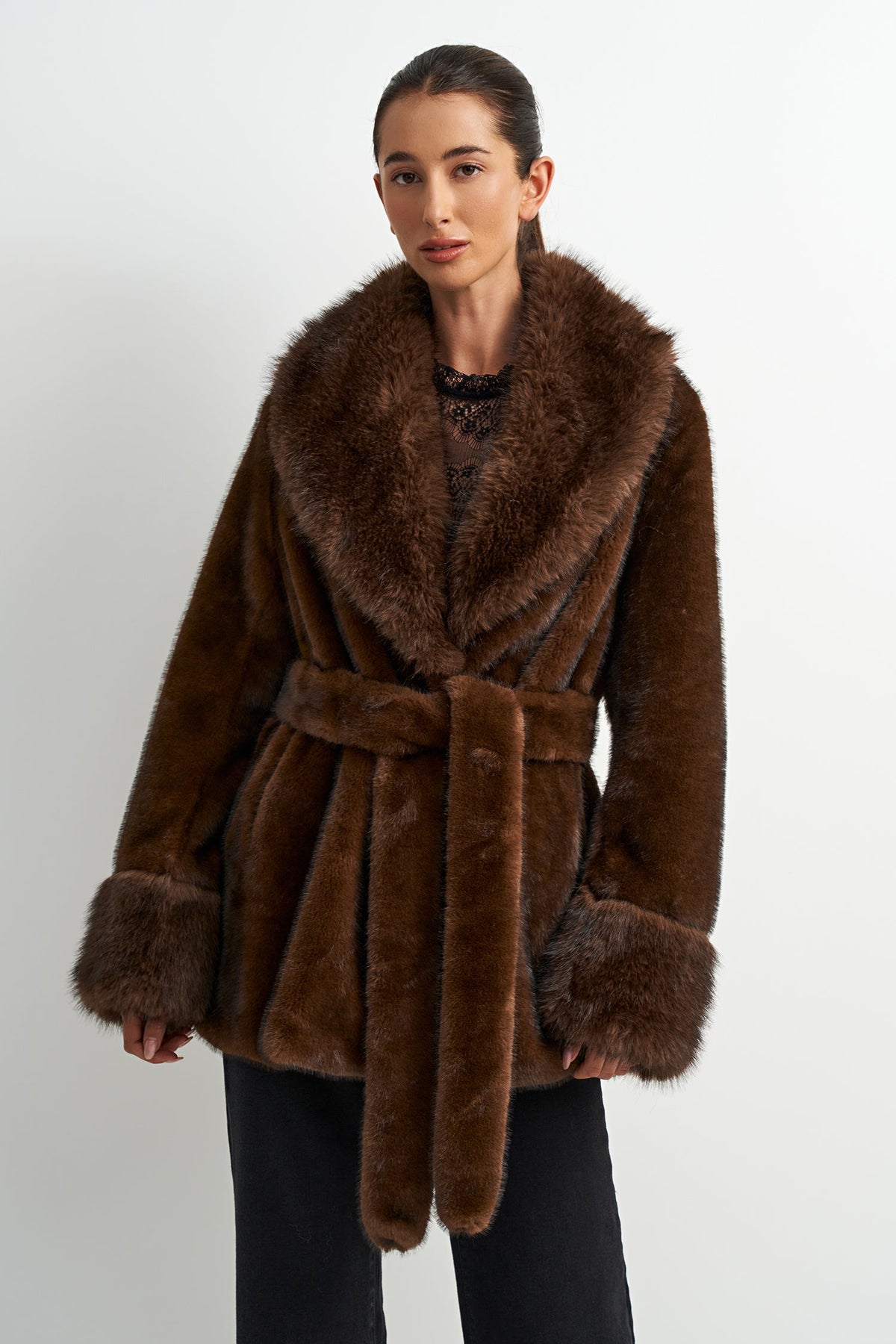 A woman modeling a brown faux fur wrap coat with a wide collar, matching belt, and ribbed cuffs, set against a plain light gray background.