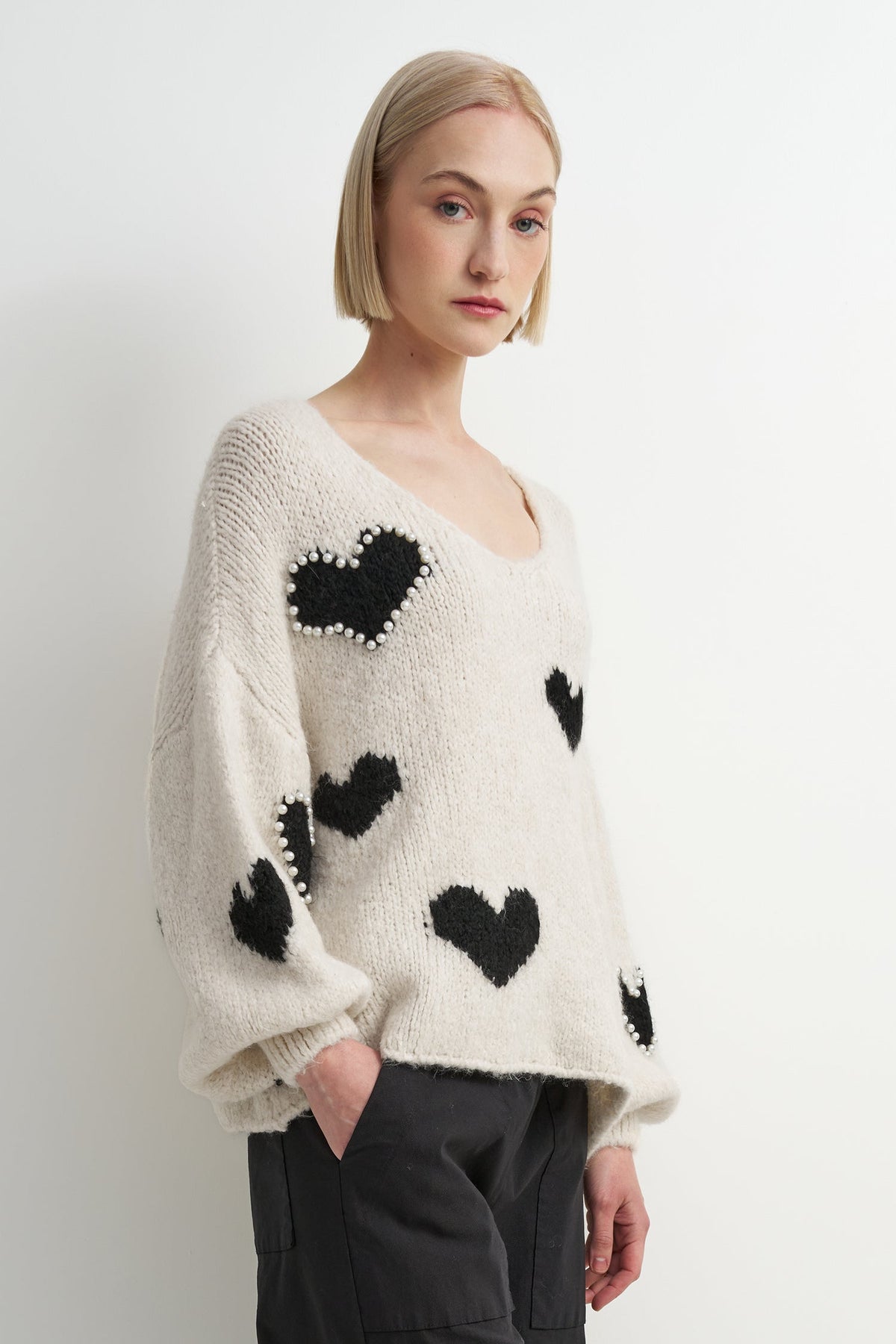 A woman wearing a cream-colored fuzzy knit sweater with black heart motifs, styled with dark gray pants against a plain light gray studio background.