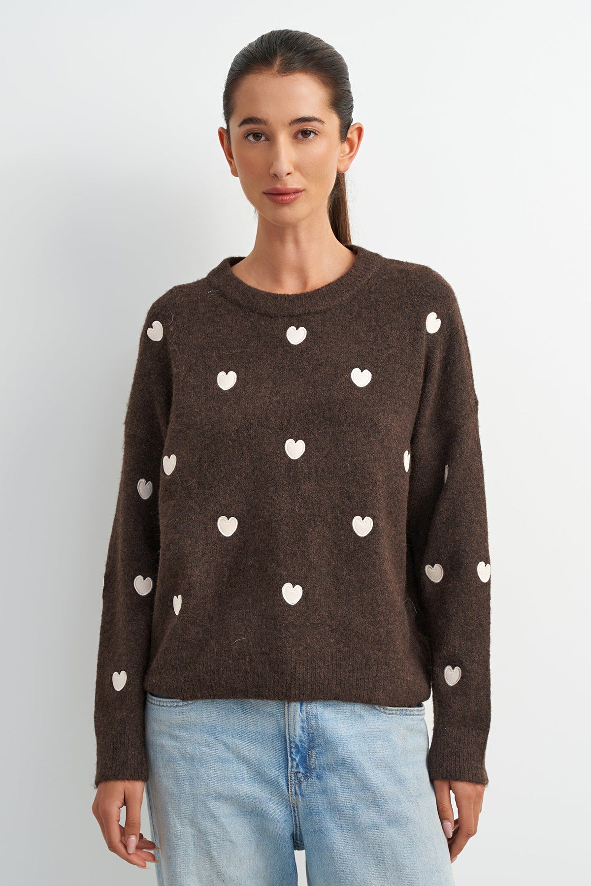 A woman wearing a dark brown knit sweater with small white heart motifs, paired with light blue jeans, standing against a light gray studio background.