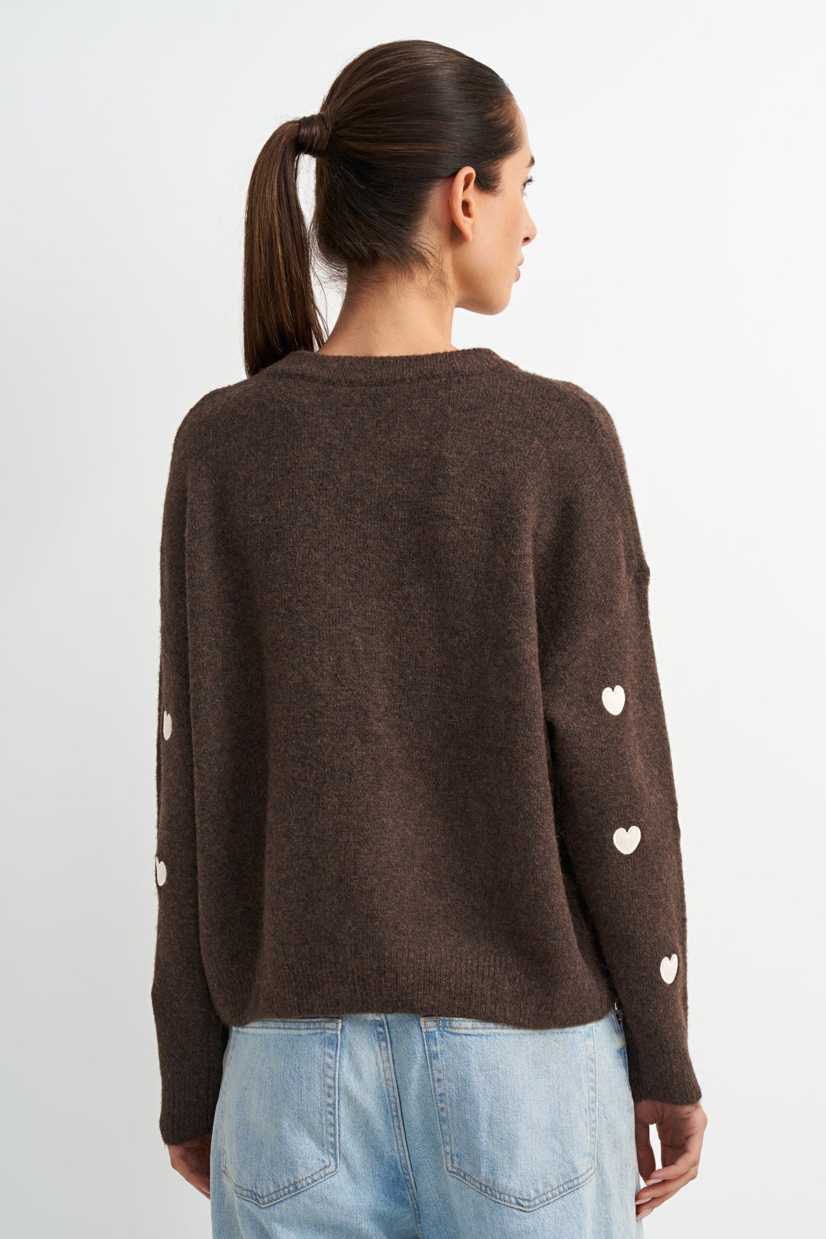 A woman shown from the back wearing a dark brown knit sweater with small white heart details on the sleeves, paired with light blue jeans, against a plain light gray background.