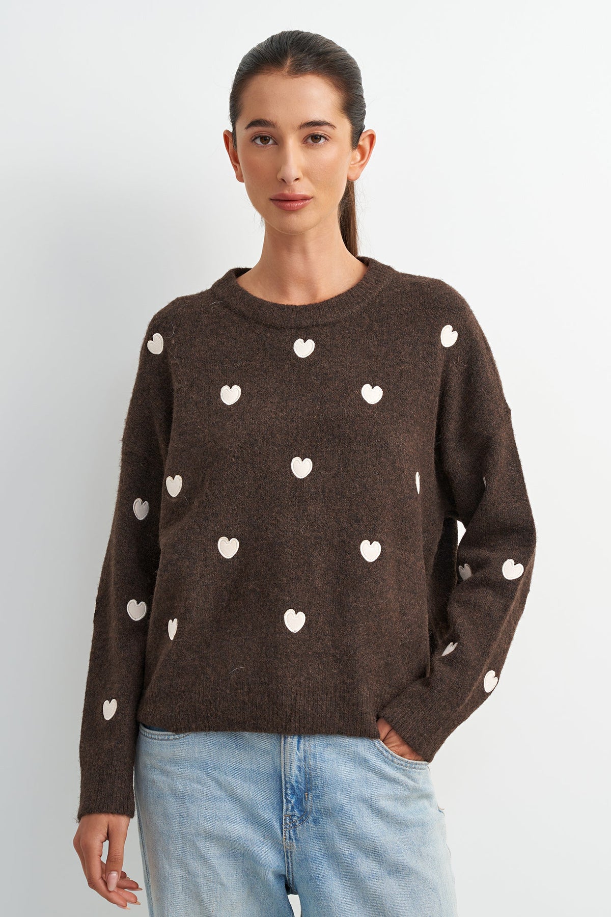 A woman wearing a dark brown knit sweater with small white heart motifs, paired with light blue jeans, standing against a plain light gray background.