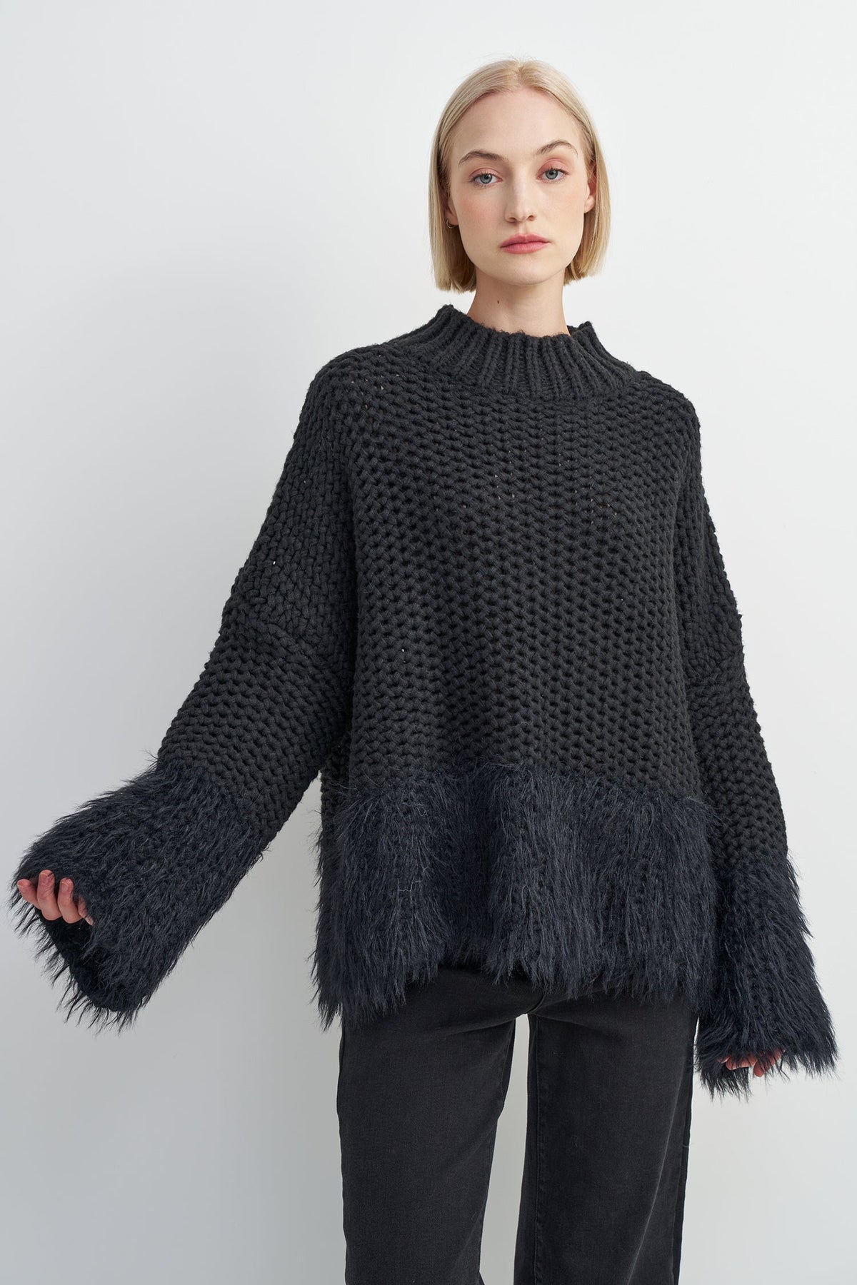 A woman modeling a dark charcoal gray chunky knit sweater with a fuzzy textured hem and wide bell sleeves, paired with black pants against a light gray studio background.