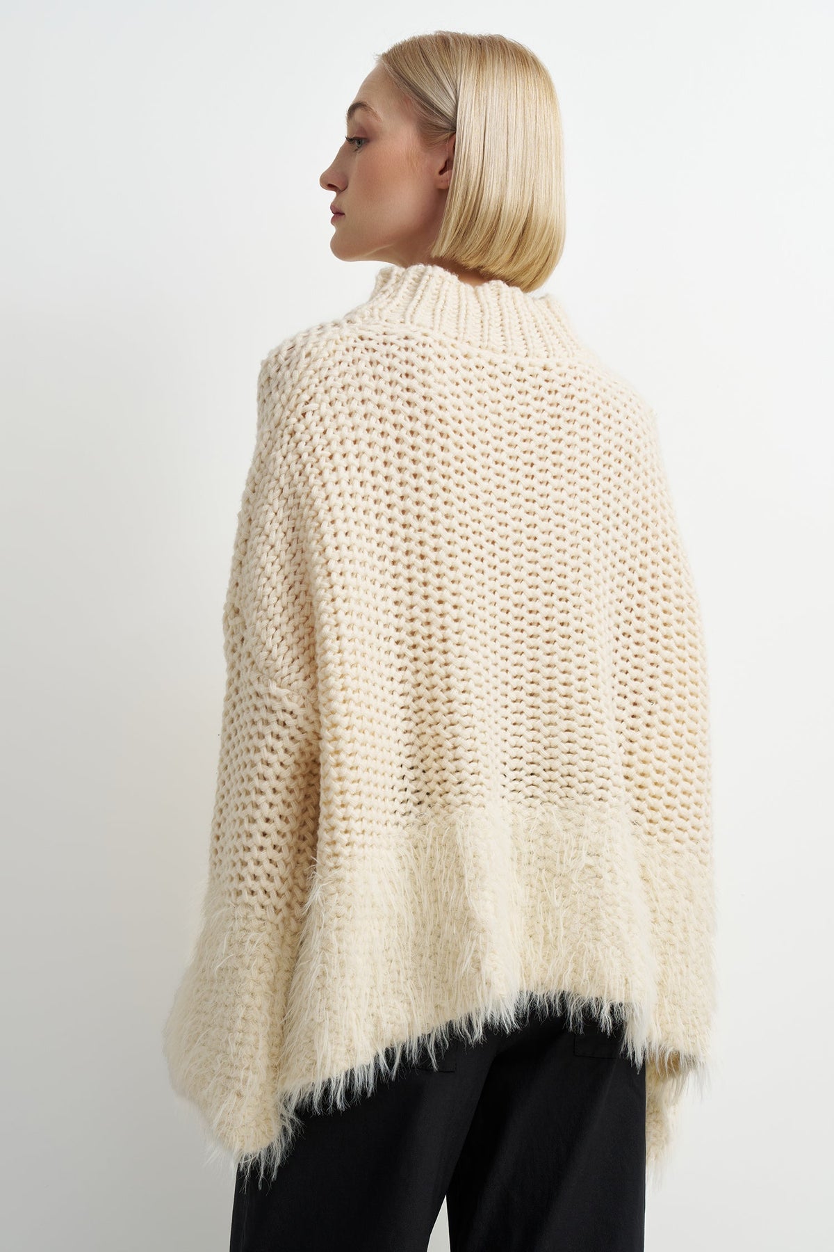 A woman shown from the back wearing a chunky knit open-knit sweater in cream, paired with black pants, against a plain light gray studio background.