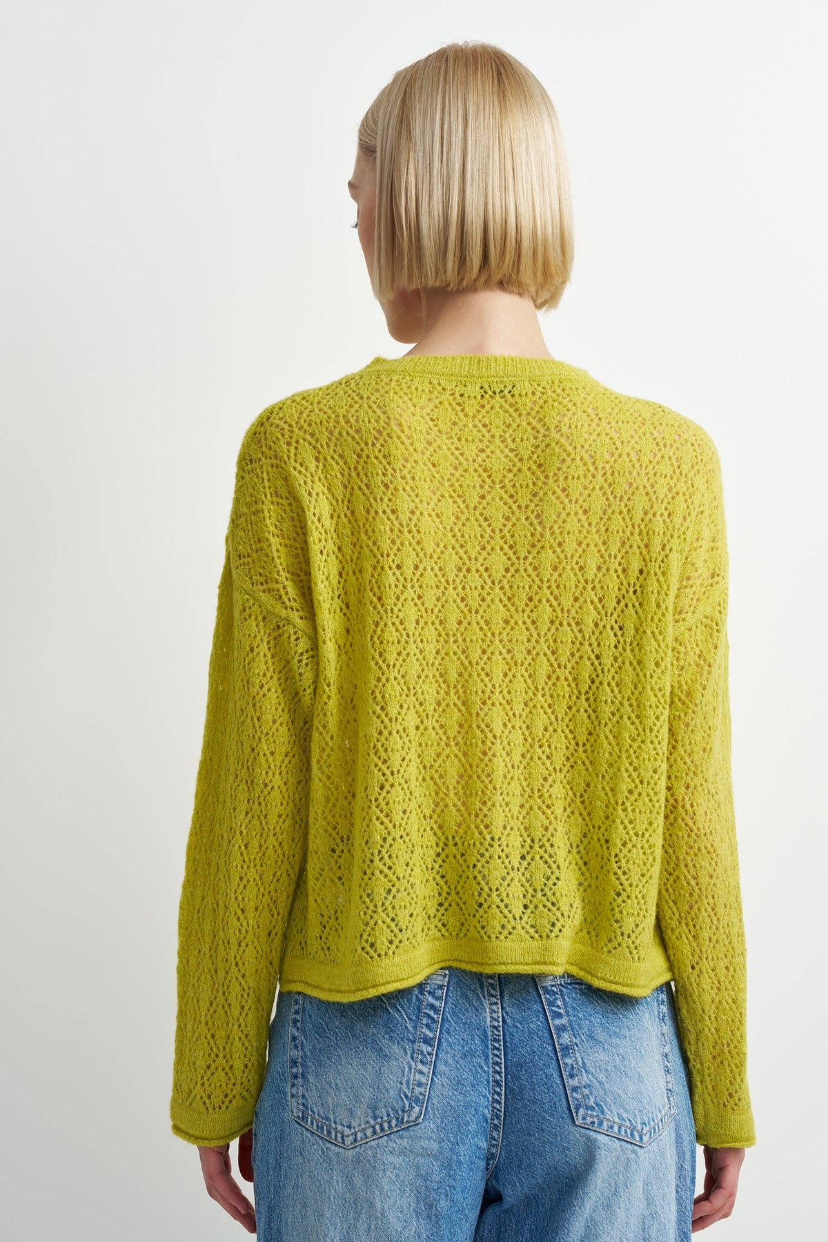 A woman viewed from the back wearing a cropped, long-sleeve knit sweater in a bright chartreuse/lime green color, paired with blue jeans, against a plain light gray studio background.