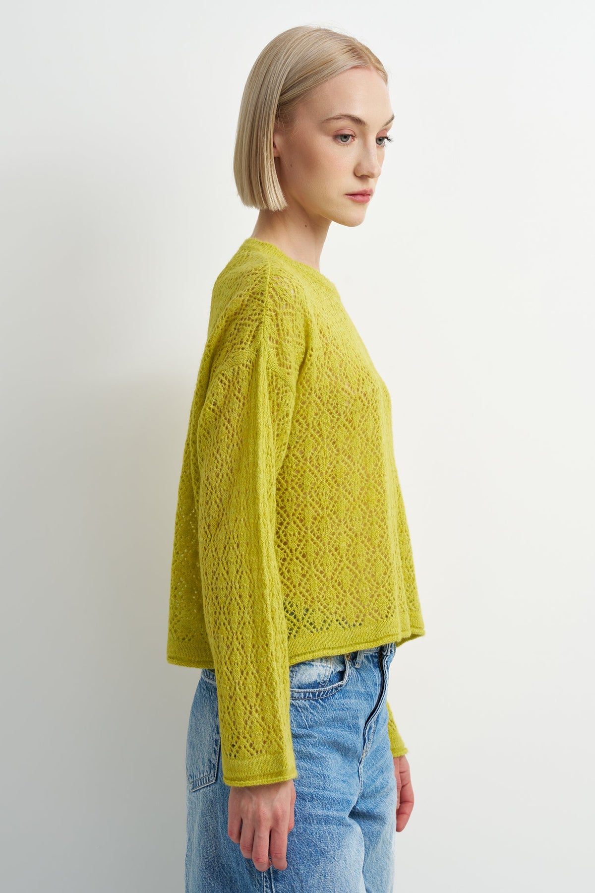 A woman with a short blonde bob is shown in profile wearing a textured, open-knit lime green sweater and light blue jeans against a plain white studio background.