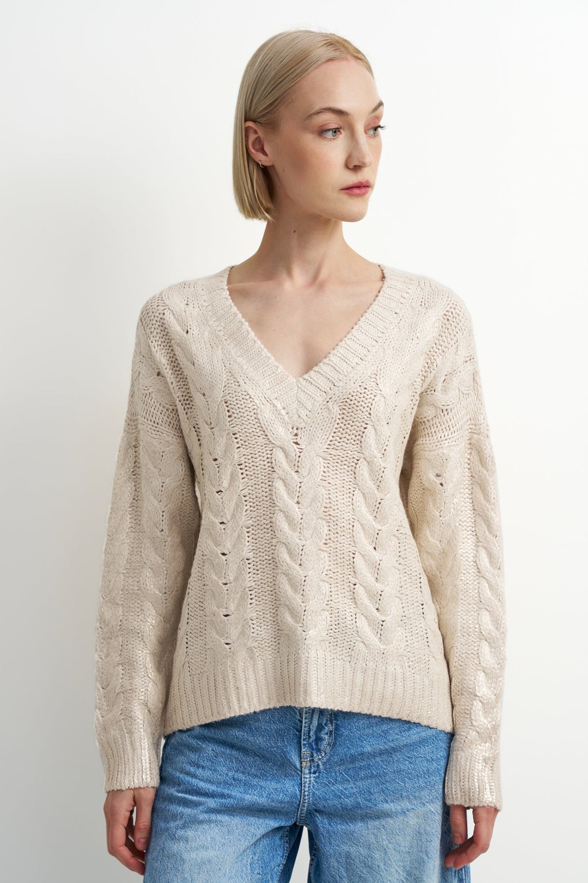 A woman wearing a cream-colored cable-knit V-neck sweater and blue jeans, photographed against a plain light gray studio background.