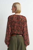 A woman shown from the back wearing a long-sleeve, gathered blouse with an abstract floral pattern in rust red, brown, and black, paired with olive green trousers against a light gray studio background.