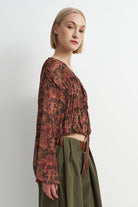 A woman shown in side profile wearing a long-sleeve, gathered wrap-style blouse with an abstract floral print in rust, brown, and burgundy tones, paired with olive green high-waisted trousers.