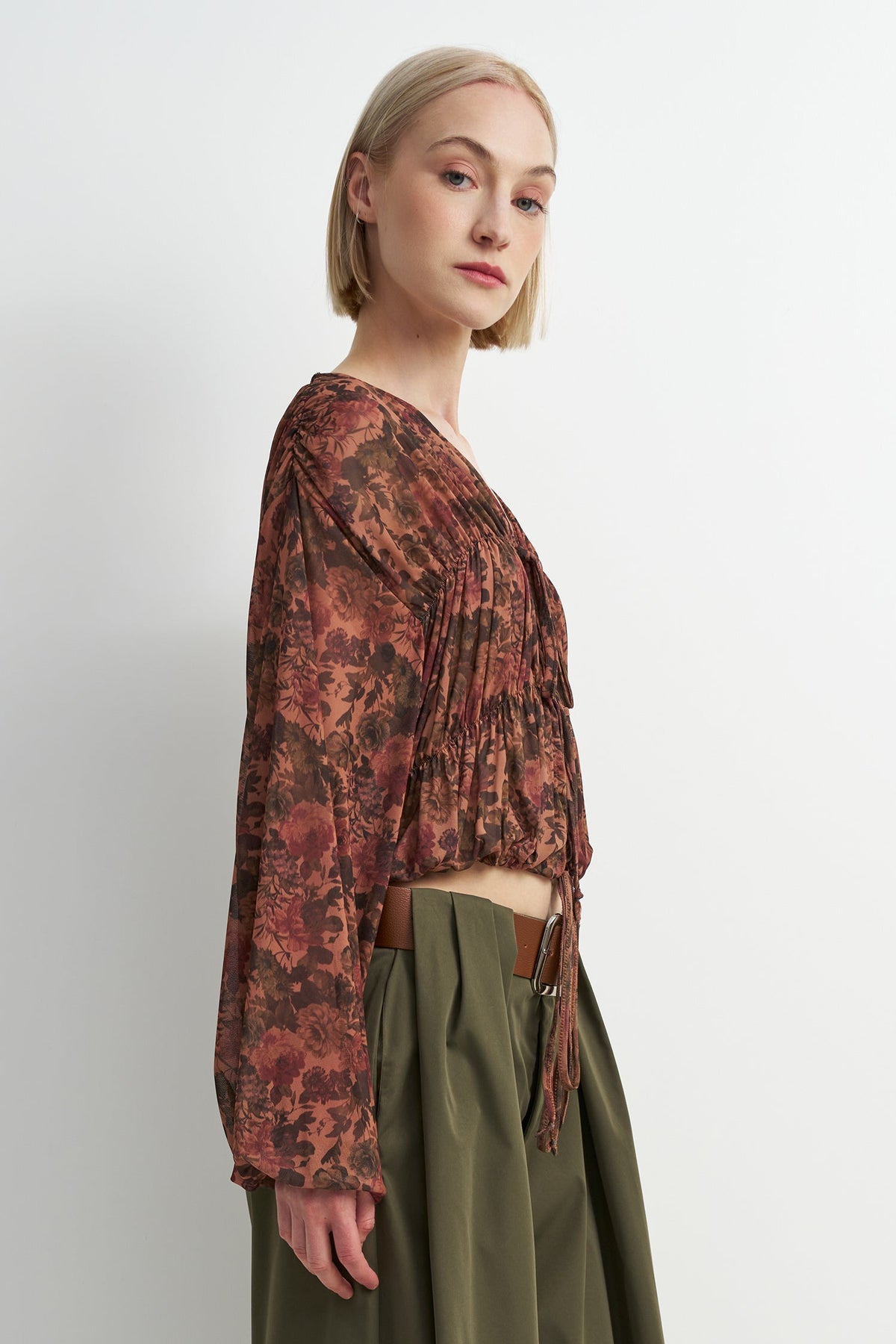 A woman shown in side profile wearing a long-sleeve, gathered wrap-style blouse with an abstract floral print in rust, brown, and burgundy tones, paired with olive green high-waisted trousers.