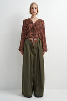 A model stands against a plain light gray studio background wearing a long-sleeve, gathered V-neck blouse with an abstract rust and brown pattern, paired with wide-leg olive green trousers and a brown belt.