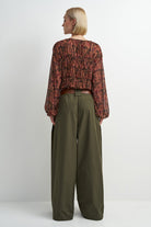A model shown from the back wearing a long-sleeve, gathered, cropped blouse with a rust-red and black floral pattern, paired with olive green wide-leg trousers, against a plain light gray background.