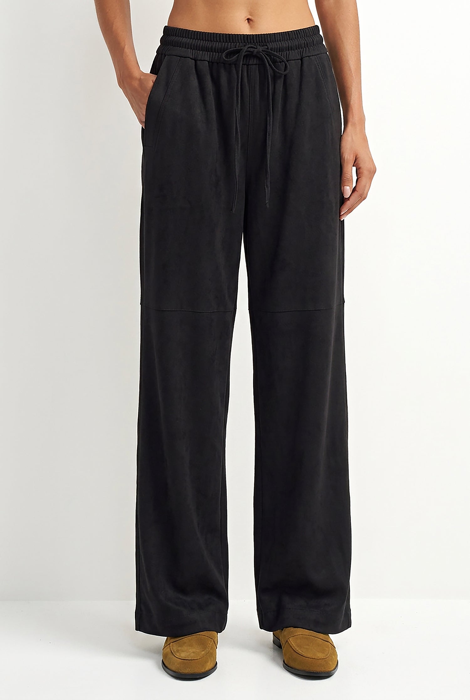 A person wearing black wide-leg drawstring pants with side pockets, paired with tan slip-on shoes, shown against a plain light gray studio background.