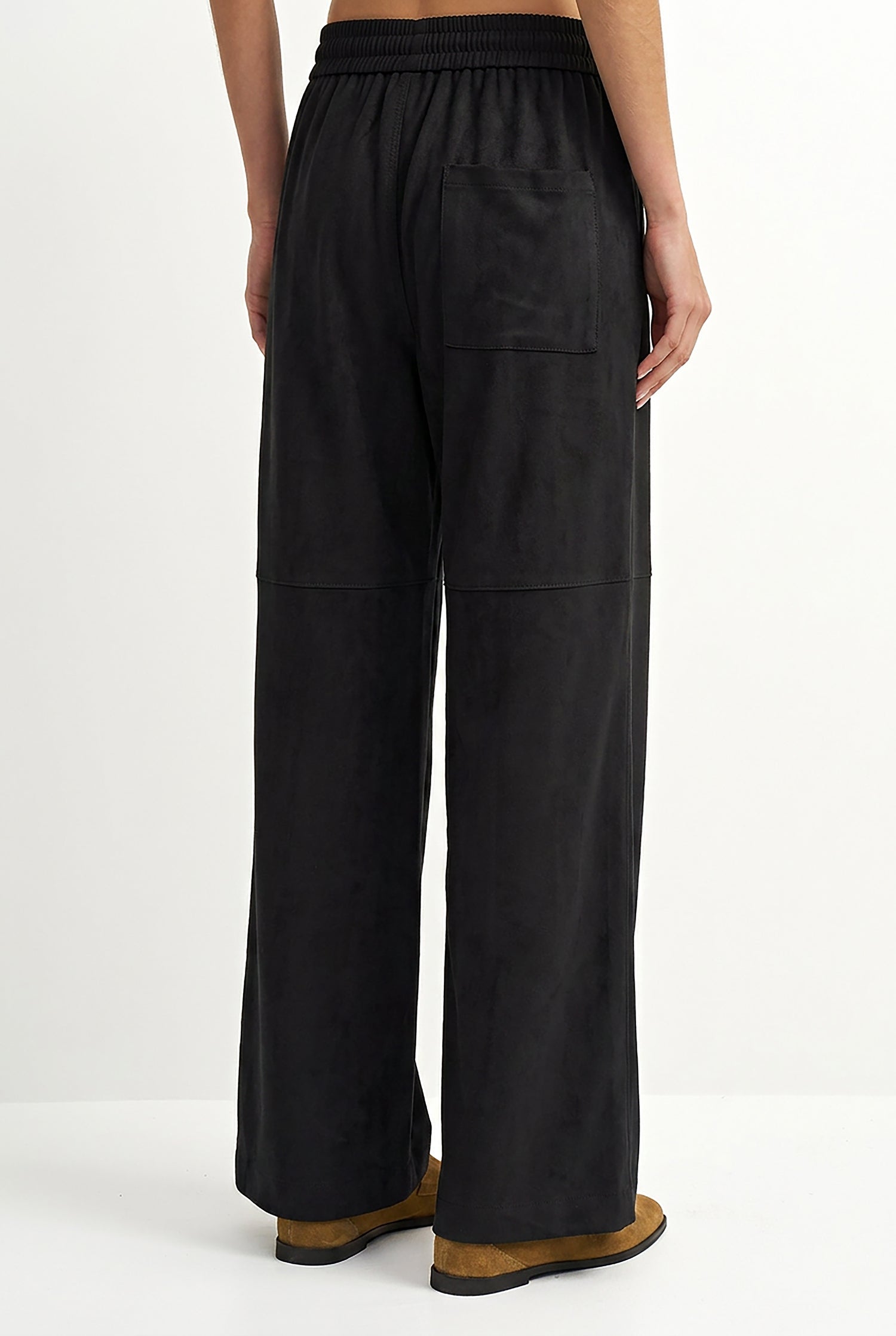 A person shown from the back wearing loose black wide-leg pants with an elastic waistband and brown shoes, photographed against a clean white studio background.