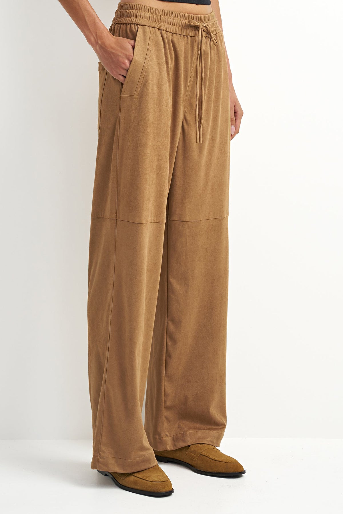 A person wearing wide-leg tan pants with an elastic waistband, drawstring, and side pockets, paired with tan shoes against a plain light background.