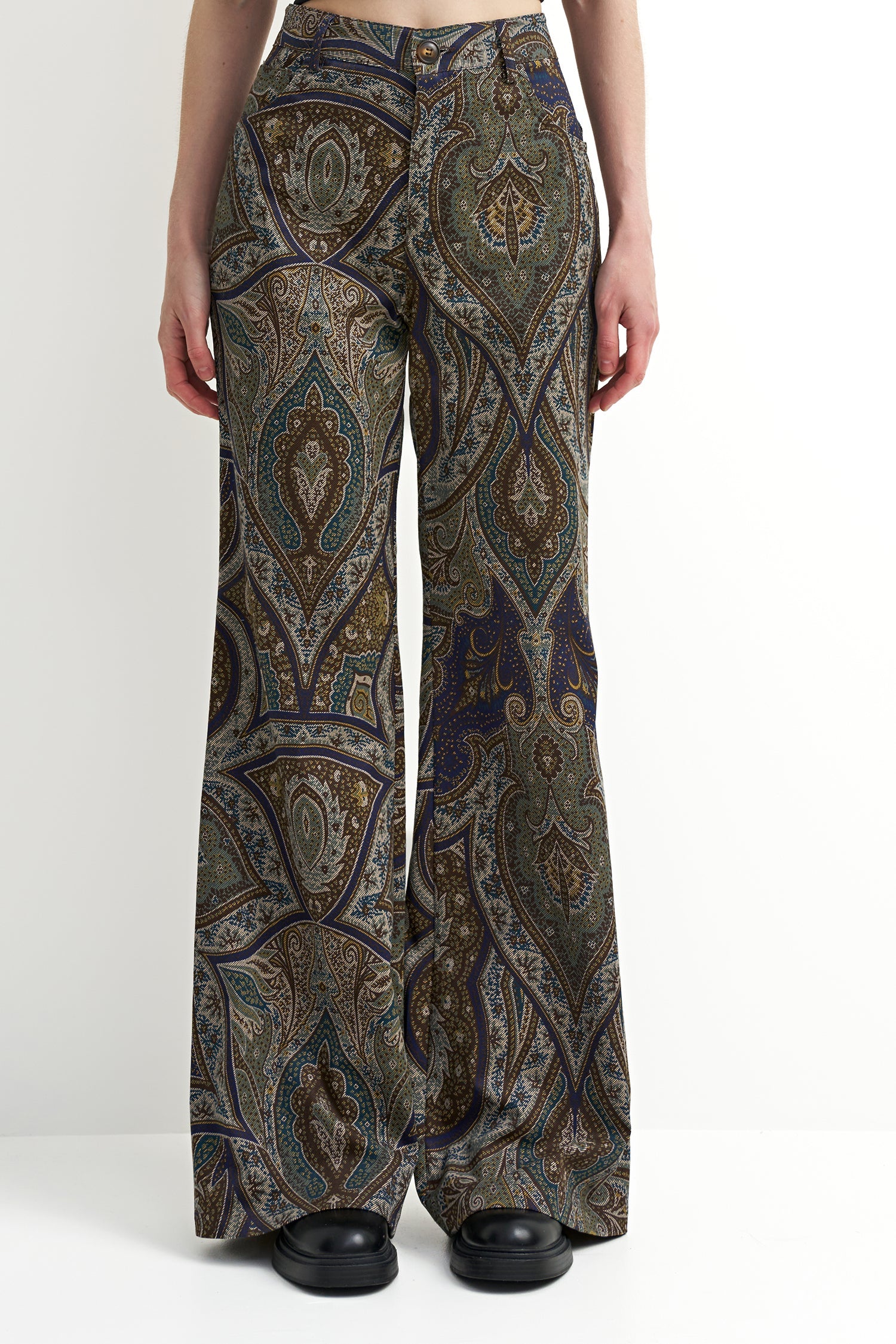 A model is shown from the waist down wearing high-waisted, wide-leg trousers with an intricate paisley pattern in olive green, brown, navy blue, and beige. The model is wearing black platform shoes against a clean white studio background.