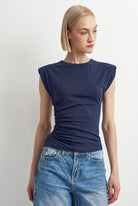 A woman modeling a navy blue sleeveless top with a ruched side detail, paired with light-wash blue jeans, against a plain light gray background.