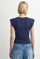 A woman with a short blonde bob is shown from the back, wearing a navy blue cap-sleeve t-shirt and light blue denim jeans against a plain light gray background.