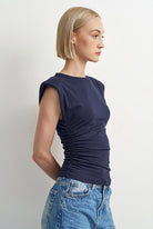 A woman with short blonde hair is shown in profile wearing a sleeveless, dark navy blue ruched top and light blue denim jeans against a plain light gray background.