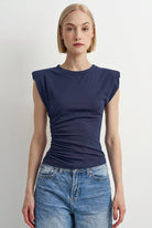 A woman modeling a navy blue sleeveless top with a ruched, gathered front and a crew neckline, paired with blue jeans, against a plain light gray background.
