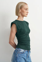 A woman with short blonde hair is shown in profile wearing a dark green ruched sleeveless top and light blue jeans against a plain light gray background.