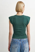A woman viewed from the back wearing a dark green sleeveless top with ruching at the back and light blue jeans, against a plain light gray studio background.