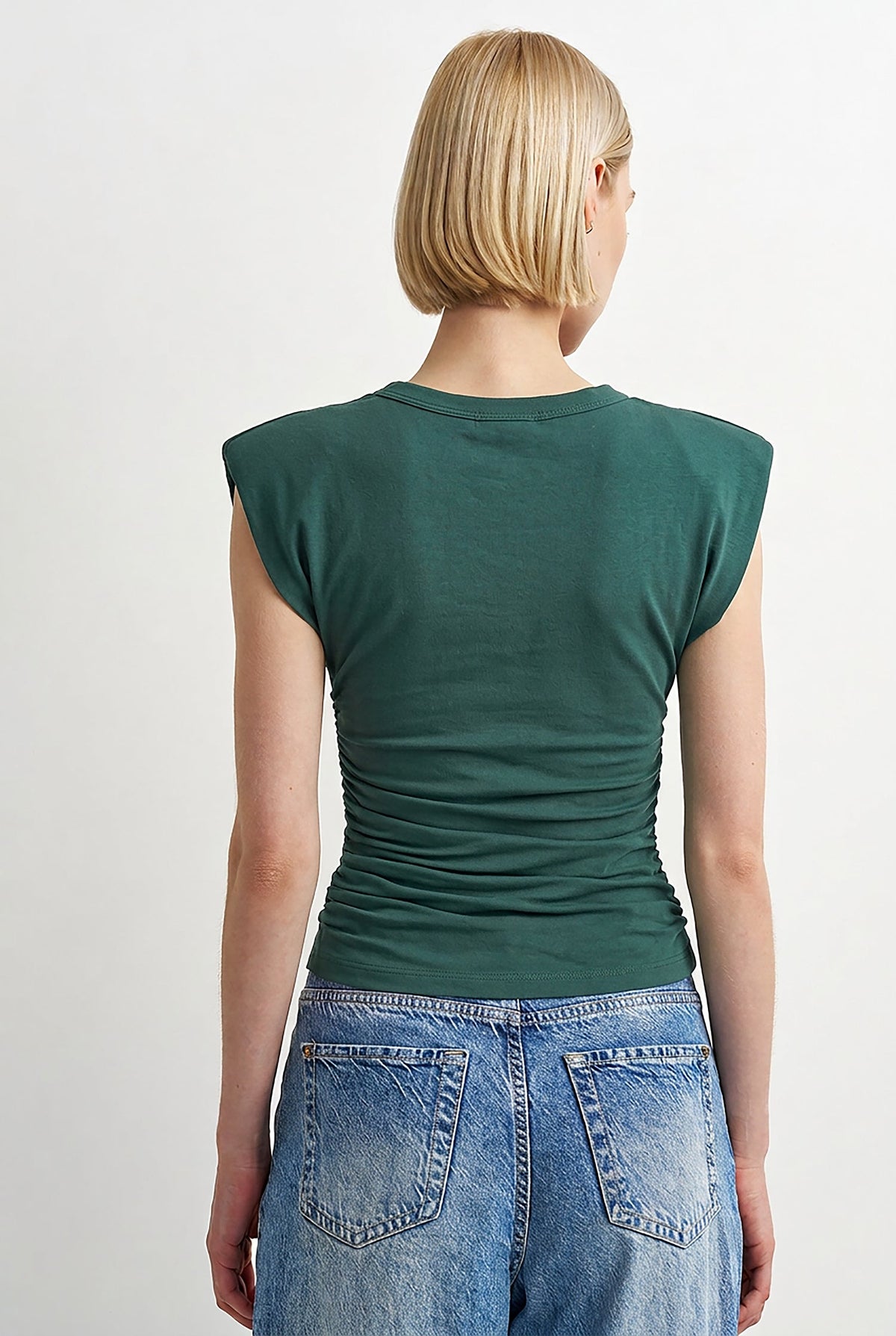 A woman viewed from the back wearing a dark green sleeveless top with ruching at the back and light blue jeans, against a plain light gray studio background.