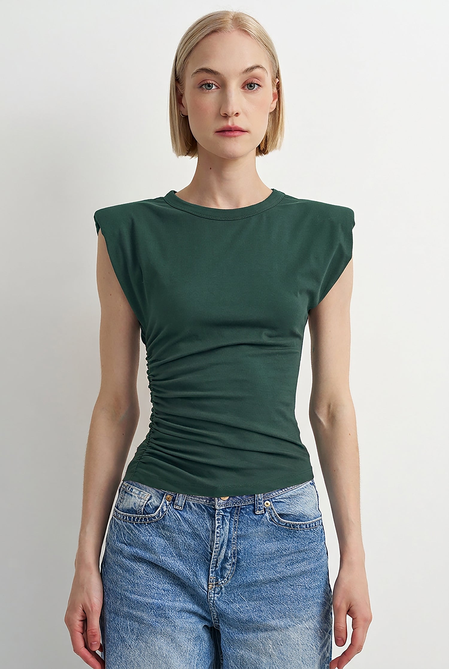 A woman modeling a dark green sleeveless ruched t-shirt with cap sleeves, paired with blue jeans, against a plain light gray background.