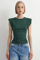 A woman modeling a dark green sleeveless ruched t-shirt with cap sleeves, paired with blue jeans, against a plain light gray background.
