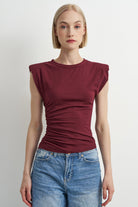 A woman modeling a sleeveless, ruched crewneck top in deep burgundy, paired with blue jeans, against a plain light gray background.
