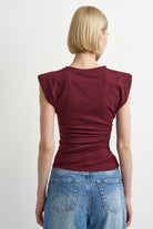 A woman with a short blonde bob is shown from the back wearing a sleeveless, ruched burgundy t-shirt and blue jeans against a plain light gray background.