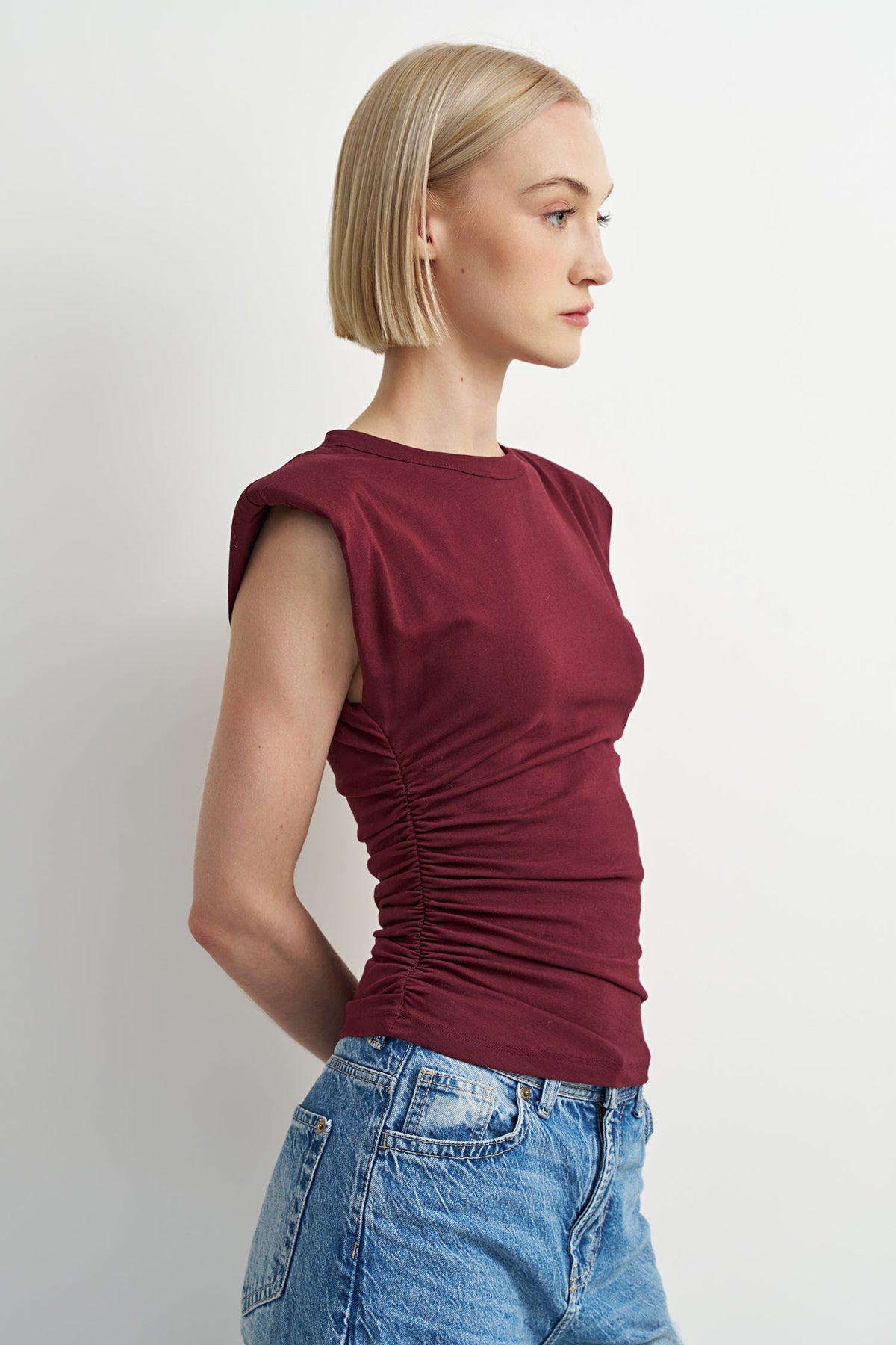 A woman with a short blonde bob is shown in profile wearing a sleeveless, ruched top in deep burgundy and blue jeans, set against a plain light gray studio background.